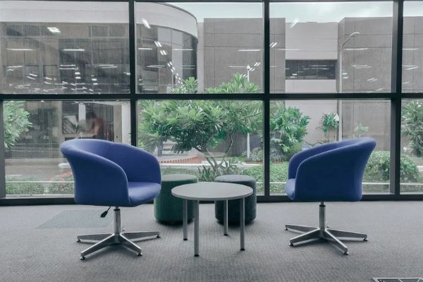 Image of an professional looking office room, with two empty blue chairs facing each other, separated by a table. Behind the table and chairs is a large window showing an outside courtyard for the building with neat trees and bushes.