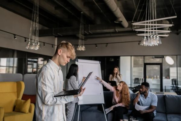 A Gen Z employee is looking concerned at something on his laptop while colleagues meet in the background, using a flip chart.